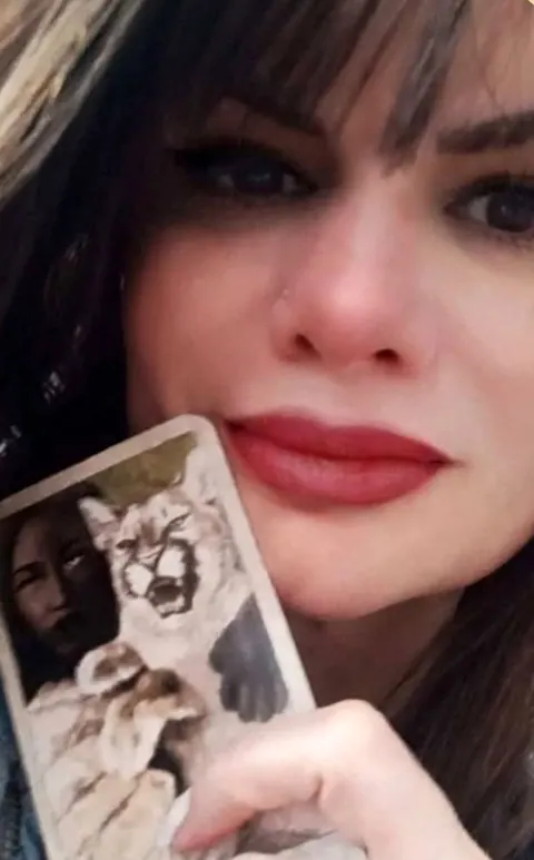 Grasiela Santos, de cabelo escuro e batom vermelho, segurando uma carta de tarô perto do rosto